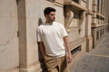 Man leaning against a stone facade in an off-white Freedom Fit Tee with beige trousers